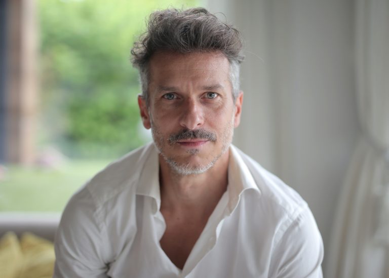 A portrait of an adult male with a quiff and blue eyes under the lights with a blurry background
