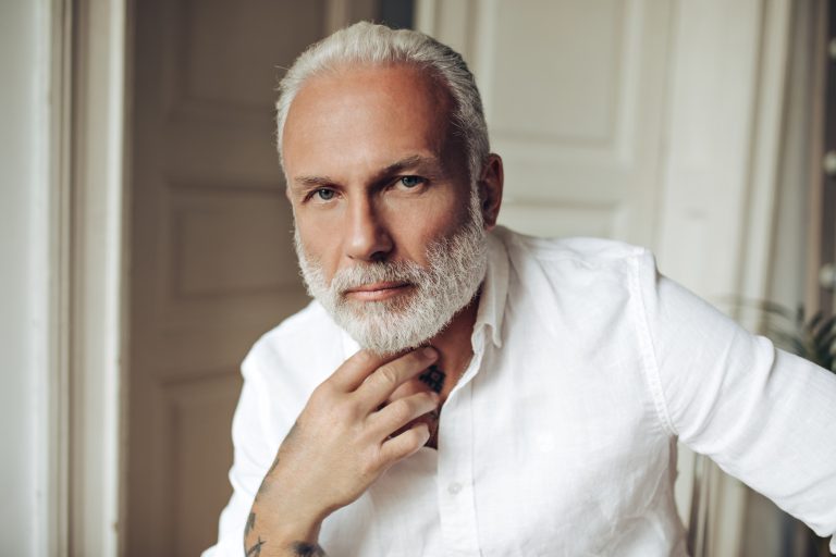 Blue eyed handsome man in white shirt looks into camera. Tattooed gray-haired adult guy with beard in bright clothes posing